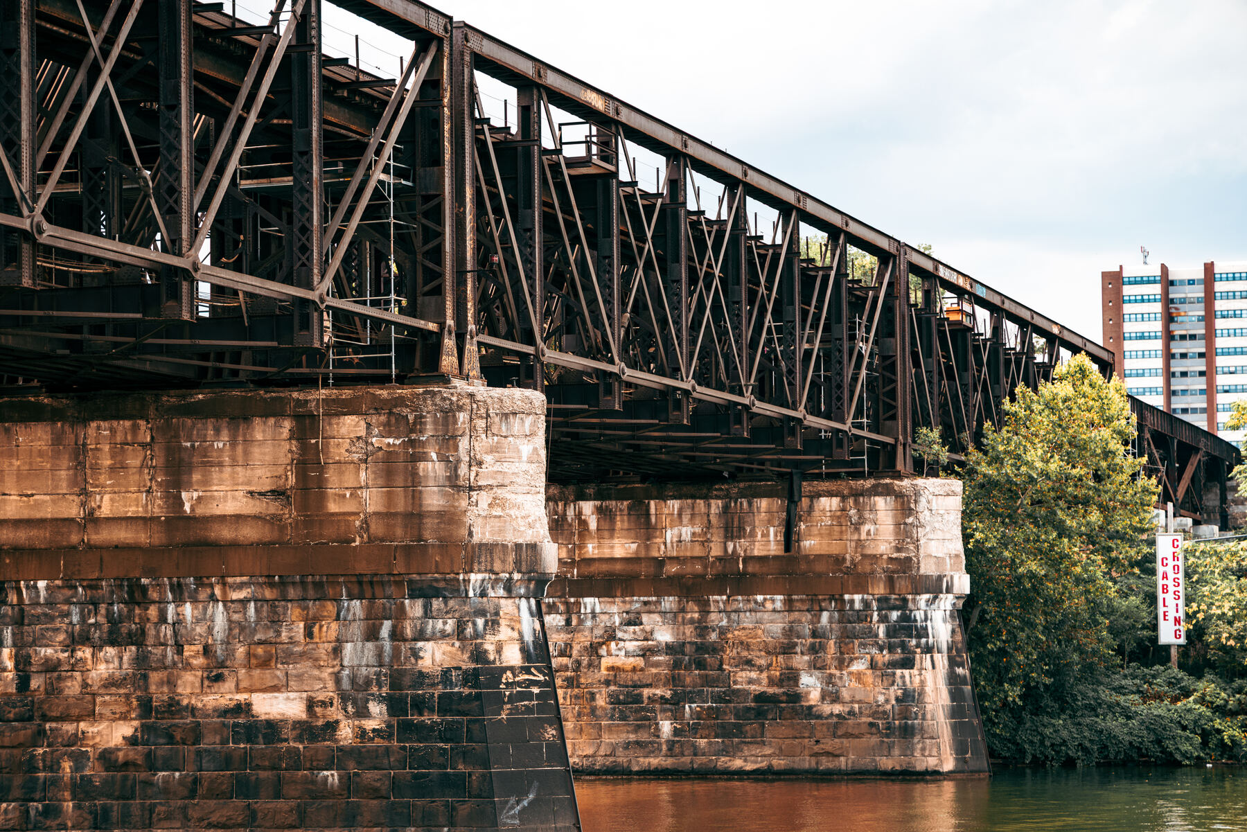 A dark steel railroad truss bridge crossing the river on stone piers